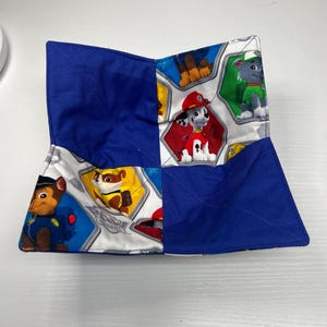 May include: A handmade fabric bowl featuring a colorful Paw Patrol design. The bowl has a blue exterior and a patterned interior with images of cartoon dogs. The bowl is designed to hold items and is made of quilted fabric.