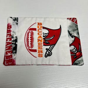 May include: A rectangular white fabric item with a red and orange embroidered football and the text "BUCCANEERS". The item features the team's logo, a skull with crossed swords, and the text "TAMPA BAY BUCCANEERS".