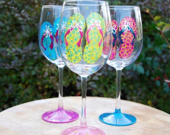 Puede incluir: Tres copas de vino con diseños de chanclas pintadas. Las copas son transparentes con bases pintadas de rosa, azul y amarillo. Las chanclas están pintadas de amarillo, rosa y azul con detalles blancos.