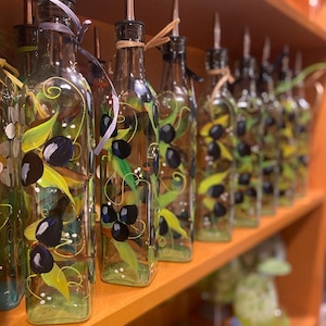 A row of beautiful hand painted olive oil bottles with olive branches wrapping around the bottles. Olive green, black, blue with highlights. whimsical vines wind between branches. a coordinating wrapphia ribbon is tied around the top. Glorious Goblets.