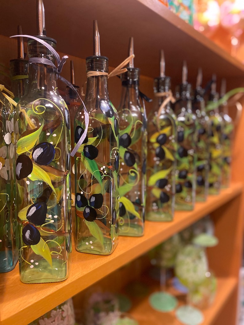 A row of beautiful hand painted olive oil bottles with olive branches wrapping around the bottles. Olive green, black, blue with highlights. whimsical vines wind between branches. a coordinating wrapphia ribbon is tied around the top. Glorious Goblets.