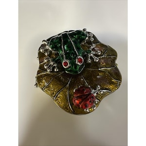 Pode incluir: Caixa de bugigangas decorativa em forma de folha, com um sapo verde e uma joaninha vermelha. O sapo tem olhos vermelhos e está em cima de uma folha com tons dourados e castanhos. A joaninha está na folha.