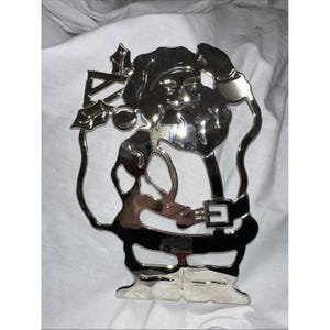 Może przedstawiać: Srebrny metalowy ornament bożonarodzeniowy w kształcie Świętego Mikołaja. Ozdoba ma wycięty wzór ze szczegółami, takimi jak pasek, broda i ostrokrzew. Jest wyświetlany na białej powierzchni.