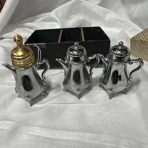 Pode incluir: Três saleiros e pimenteiros prateados em miniatura em forma de bule. Um dos saleiros tem uma tampa dourada. Os saleiros têm pegas e assentam em três pés pequenos. Uma caixa preta com três compartimentos está no fundo.