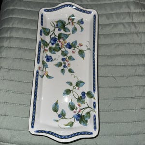 Puede incluir: Bandeja rectangular de cerámica blanca con un diseño floral azul y blanco. La bandeja presenta un borde de volutas azules y está decorada con arándanos, hojas y pequeñas flores rosas.
