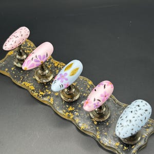 Può includere: Esposizione di cinque unghie finte, ciascuna montata su un supporto argentato. Le unghie sono di forma ovale e dipinte in vari colori pastello, tra cui rosa e blu, con disegni floreali e maculati. L'esposizione è su un vassoio trasparente con accenti di foglia d'oro.
