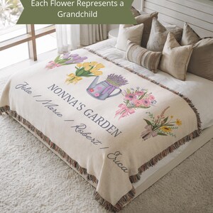 May include: Cream-colored throw blanket with a floral design and the text "NONNA'S GARDEN". The blanket features illustrations of flowers, a watering can, and the names "Jolie / Marie / Robert / Erica". The blanket has a fringed edge.