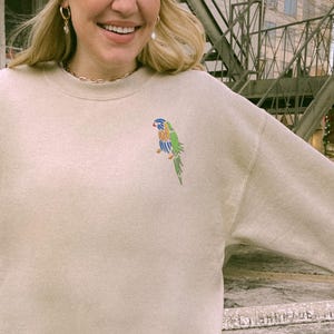 May include: A beige sweatshirt with a colorful parrot embroidered on the front.