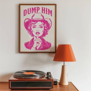 Pode incluir: Uma impressão emoldurada com uma ilustração rosa de uma mulher com um chapéu de cowboy, com o texto "DUMP HIM". Uma vitrola e uma lâmpada laranja estão em primeiro plano.