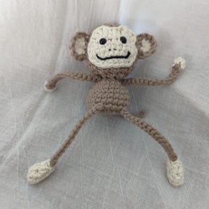 May include: A handmade crochet monkey doll in shades of brown and cream. The monkey has a light brown body, with a cream-colored face, hands, and feet. The monkey's face features black eyes and a black smile. The arms and legs are outstretched.