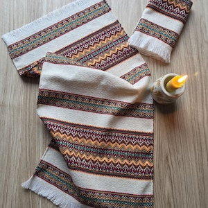 May include: A cream-colored table runner and two matching towels with a geometric pattern in burgundy, teal, and gold. The runner and towels have fringed edges. A lit candle sits in a small ceramic holder.