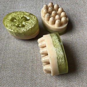 Op de afbeelding: Drie handgemaakte zepen met natuurlijke luffa en massagefuncties. Eén zeep is ovaal van vorm met een luffa-kern. Een andere heeft een afgeronde bovenkant met massagenoppen. De derde zeep heeft een luffa-kern en massagenoppen.