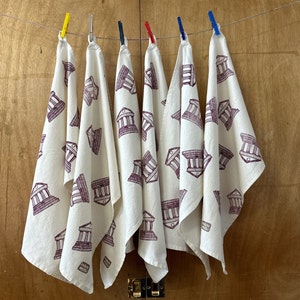 May include: Five white tea towels with a burgundy print of a building with columns. The towels are hanging on a clothesline with clothespins.