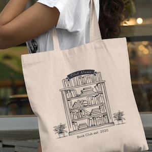 Peut inclure: Sac fourre-tout beige avec une illustration en noir et blanc d'une bibliothèque remplie de livres. Le texte "Club Name" est au-dessus de la bibliothèque et "Book Club est. 2020" en dessous. Deux plantes en pot encadrent le dessin.