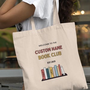 Peut inclure: Sac fourre-tout crème avec le texte "WELCOME TO THE CUSTOM NAME BOOK CLUB EST.2025". En dessous, un graphisme de livres colorés sur une étagère. Le sac est tenu par une personne.