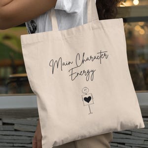 Peut inclure: Un sac fourre-tout beige avec du texte noir qui dit "Main Character Energy". Un simple bonhomme bâton avec un cœur est imprimé sous le texte.