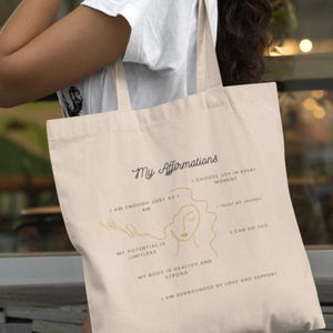 Peut inclure: Un sac fourre-tout beige avec le texte "My Affirmations" imprimé en noir. Le sac présente un dessin au trait d'un visage de femme avec des affirmations écrites autour du dessin. Les affirmations incluent "I am enough just as I am", "Choose joy in every moment", "My potential is limitless", "My body is healthy and strong", "Trust my journey", "I can do this", et "I am surrounded by love and support."