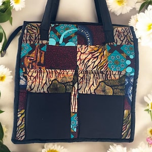 Op de afbeelding: Een patchwork tote bag met een zwarte basis en handvatten. De tas heeft stoffen met patronen in teal, bruin en geel. Twee grote zwarte zakken zitten aan de voorkant. Witte bloemen omringen de tas.