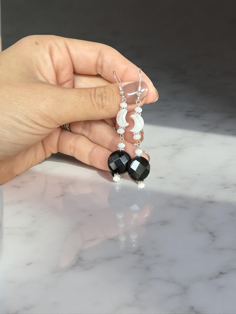 Puede incluir: Pendientes colgantes con cuentas negras facetadas y dijes de media luna blancos. Los pendientes tienen ganchos plateados y peque&ntilde;as cuentas blancas. El dise&ntilde;o es elegante y celestial.
