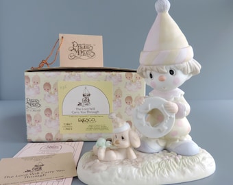 May include: A porcelain figurine of a clown wearing a pastel hat and polka dot outfit, holding a white ring, accompanied by a small dog with a turtle. The box is labeled Precious Moments, The Lord Will Carry You Through, 12467, 1 piece.