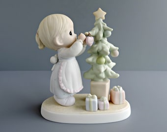 May include: A porcelain figurine of a child with blonde hair wearing a light blue dress with a pink-trimmed apron, placing a pink heart-shaped ornament labeled Good Love onto a pale green Christmas tree topped with a yellow star.