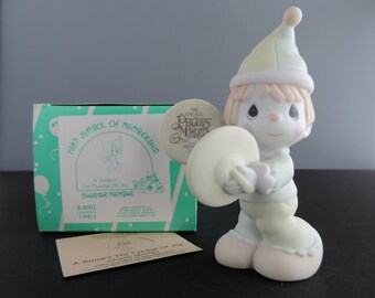 May include: A porcelain figurine of a child wearing a pastel green and yellow jester hat and outfit, holding two cymbals. The figurine is accompanied by a teal and white box labeled 1987 Symbol of Membership and a small paper card with matching text.