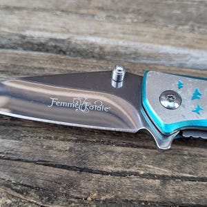 May include: A blue pocket knife with a silver blade and a butterfly design. The blade is engraved with the words "Femme Fatale".