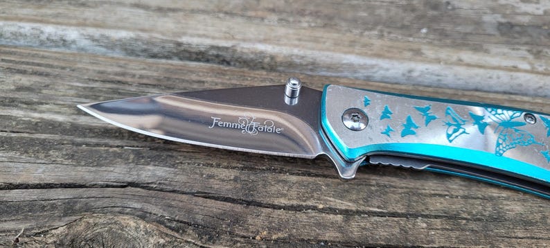 May include: A blue pocket knife with a silver blade and a butterfly design. The blade is engraved with the words "Femme Fatale".