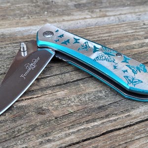 May include: A teal pocket knife with a butterfly design engraved on the handle. The blade is partially open and the knife is on a wooden surface.