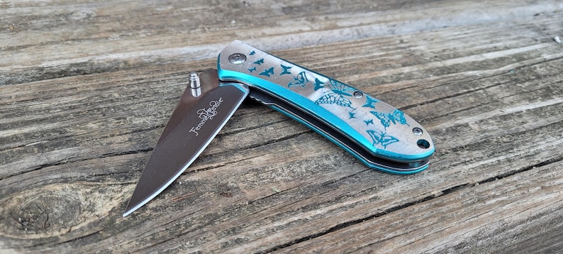 May include: A teal pocket knife with a butterfly design engraved on the handle. The blade is partially open and the knife is on a wooden surface.