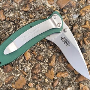 Puede incluir: Una navaja verde con un clip de bolsillo plateado y una hoja parcialmente expuesta. La hoja es plateada y tiene el texto "1520GRN, KAI, Patented, MADE IN USA". La navaja est&aacute; sobre un lecho de peque&ntilde;as rocas.