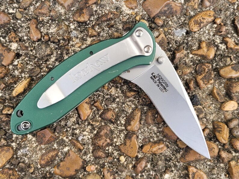 Puede incluir: Una navaja verde con un clip de bolsillo plateado y una hoja parcialmente expuesta. La hoja es plateada y tiene el texto "1520GRN, KAI, Patented, MADE IN USA". La navaja est&aacute; sobre un lecho de peque&ntilde;as rocas.
