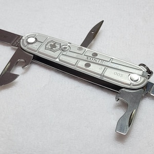 May include: A silver multi-tool with multiple blades and tools extended. The tool has a silver handle with the word "Rinox" and the number "003" printed on it. The blades are silver and the tool is on a white surface.