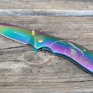 Può includere: Un coltello pieghevole arcobaleno con un design a forma di teschio inciso sul manico. La lama è parzialmente aperta e il coltello è su una superficie di legno. Il coltello ha la scritta "WARTECH" stampata sulla lama.