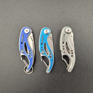 Pode incluir: Três pequenas facas de bolso dobráveis com lâminas prateadas e cabos coloridos. Duas facas têm cabos azuis e uma tem um cabo prateado. Cada faca tem um pequeno mosquetão e um porta-chaves.