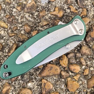 Puede incluir: Una navaja de bolsillo plegable verde y plateada con la marca "Kershaw" visible en la hoja. La navaja tiene un clip de bolsillo plateado y una hoja parcialmente expuesta. El mango es de color verde liso.
