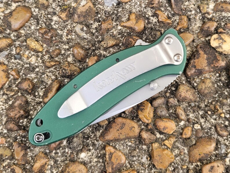 Puede incluir: Una navaja de bolsillo plegable verde y plateada con la marca "Kershaw" visible en la hoja. La navaja tiene un clip de bolsillo plateado y una hoja parcialmente expuesta. El mango es de color verde liso.