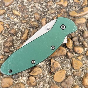 Puede incluir: Una navaja de bolsillo plegable verde con una hoja plateada. El cuchillo tiene un mango verde texturizado con tres tornillos visibles y un pivote plateado. El cuchillo est&aacute; sobre una superficie de peque&ntilde;as rocas y guijarros.