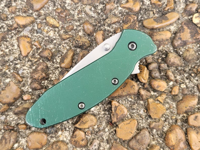 Puede incluir: Una navaja de bolsillo plegable verde con una hoja plateada. El cuchillo tiene un mango verde texturizado con tres tornillos visibles y un pivote plateado. El cuchillo est&aacute; sobre una superficie de peque&ntilde;as rocas y guijarros.