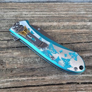 May include: A teal pocket knife with a butterfly design. The knife has a silver blade and a gold butterfly accent.