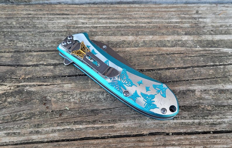 May include: A teal pocket knife with a butterfly design. The knife has a silver blade and a gold butterfly accent.