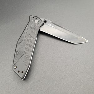 May include: Black folding knife with a partially open blade. The handle has a textured design with three visible screws. The blade is silver and has the text "1990 KAI Patented".