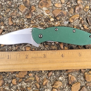 Puede incluir: Una navaja plegable con una hoja plateada y un mango verde. El cuchillo est&aacute; junto a una regla de madera, que mide aproximadamente 15 cm de largo. El cuchillo est&aacute; sobre una superficie de peque&ntilde;as rocas y guijarros.