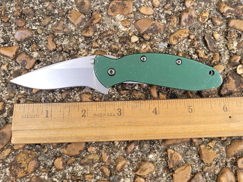 Puede incluir: Una navaja plegable con una hoja plateada y un mango verde. El cuchillo est&aacute; junto a una regla de madera, que mide aproximadamente 15 cm de largo. El cuchillo est&aacute; sobre una superficie de peque&ntilde;as rocas y guijarros.