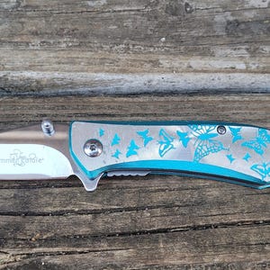 May include: A teal blue pocket knife with a silver blade and a butterfly design. The knife has the words "Female Tribe" engraved on the blade.