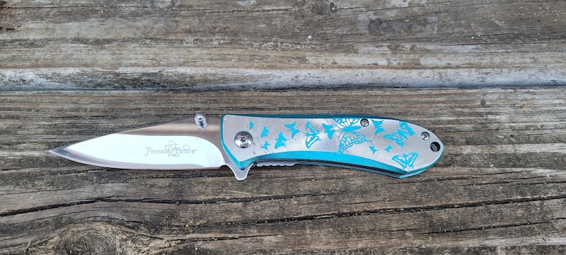 May include: A teal blue pocket knife with a silver blade and a butterfly design. The knife has the words "Female Tribe" engraved on the blade.