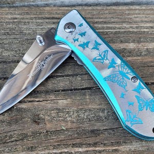May include: A silver pocket knife with a blue handle and a butterfly design. The knife is open and the blade is visible. The text "Titanium Blade" is engraved on the blade.