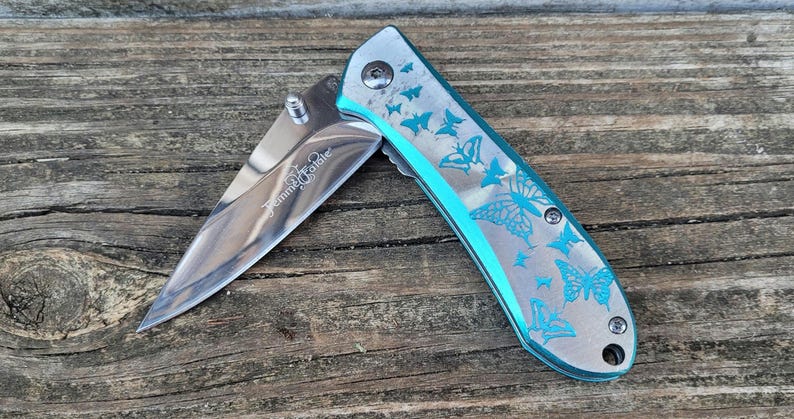 May include: A silver pocket knife with a blue handle and a butterfly design. The knife is open and the blade is visible. The text "Titanium Blade" is engraved on the blade.