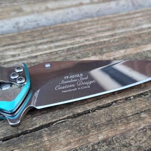 May include: A turquoise pocket knife with a butterfly design on the handle. The blade is open and the inscription reads "FF-A010LB Stainless Steel Custom Design Handcraft in China."