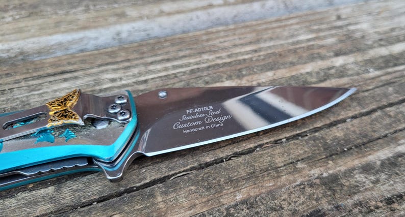 May include: A turquoise pocket knife with a butterfly design on the handle. The blade is open and the inscription reads "FF-A010LB Stainless Steel Custom Design Handcraft in China."